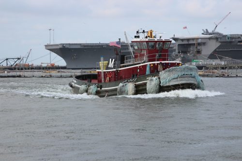 Red tugboat with fendered bow passing an aircraft carrier